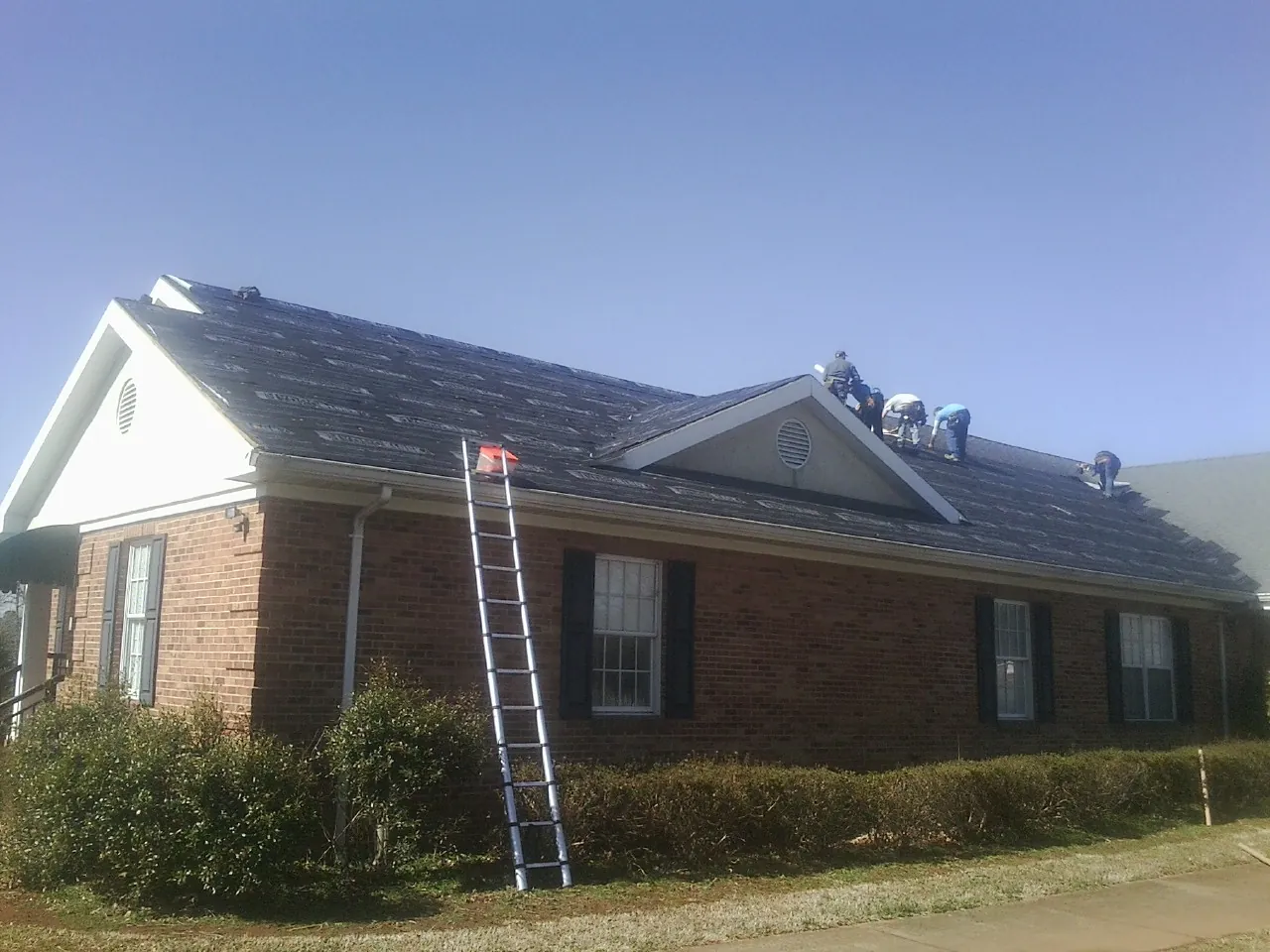Crew tearing shingles off a brick ranch roof with a ladder set — Emergency Roof Repair work by our crew in Fort Lawn