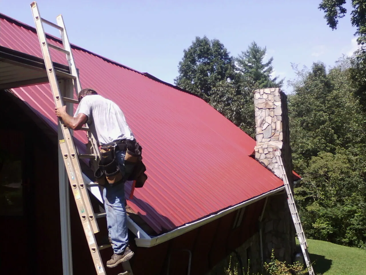 Roofer on a ladder beside a red metal roof and stone chimney — Soffit & Fascia Repair by Roofing Brothers in Fort Lawn